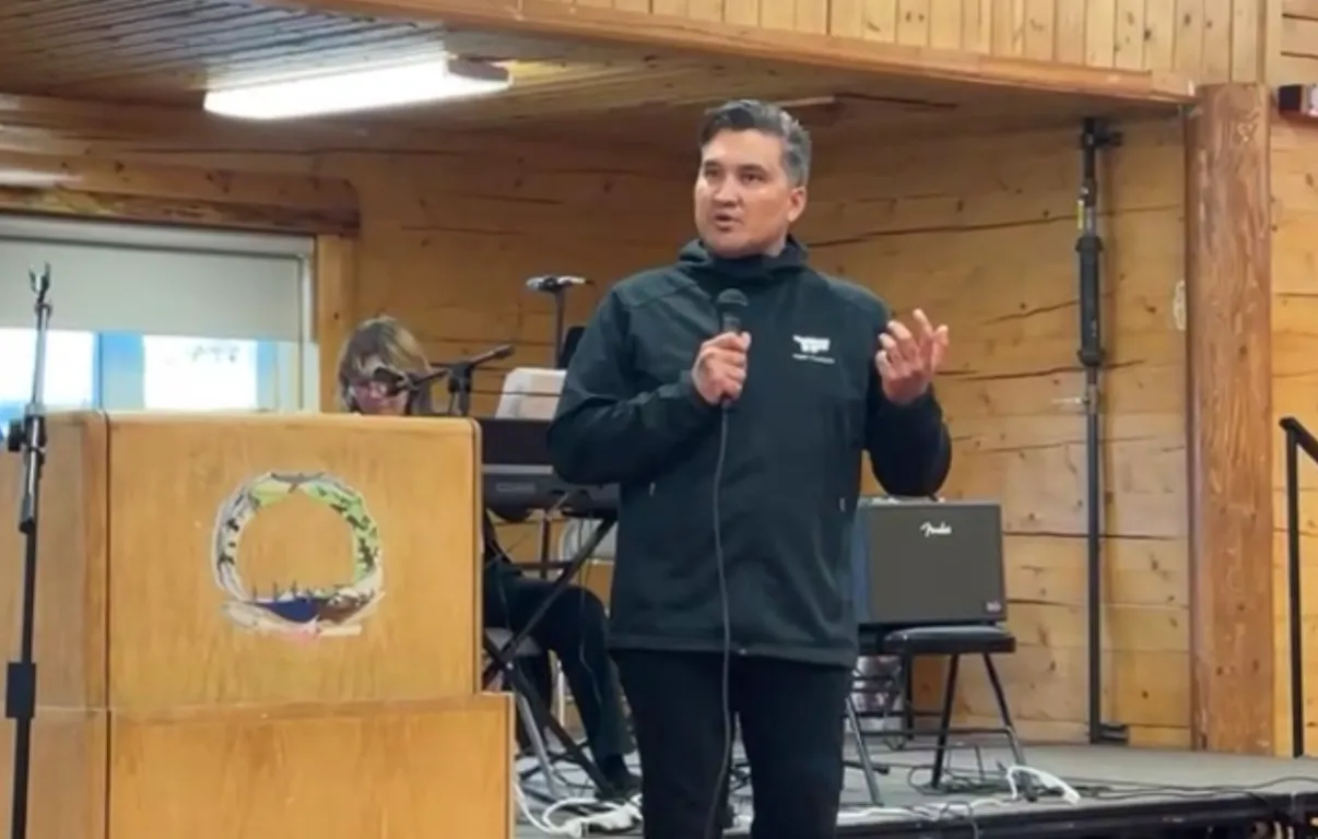 Minister Tommy Frankson in a black TF Ministries jacket preaching from a log-cabin chapel pulpit in rural Alaska.